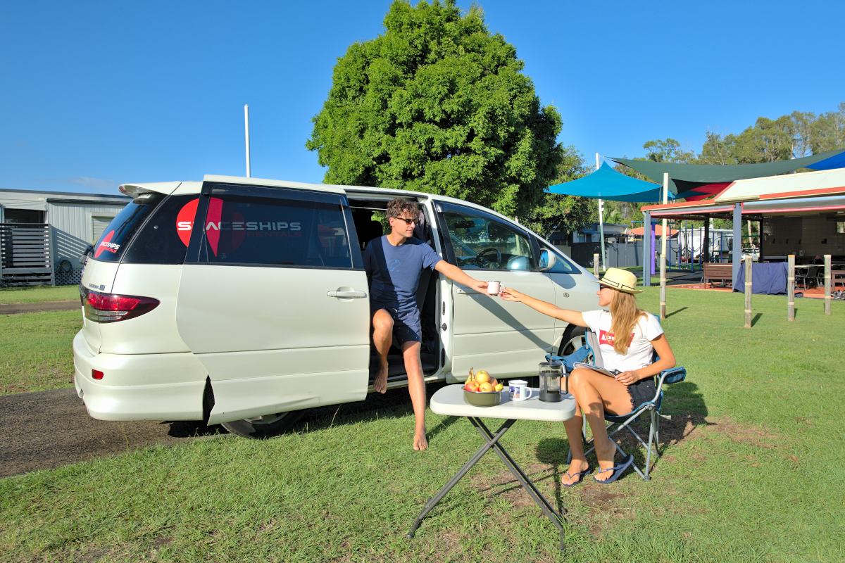 Campervan parked at a campgroun with a man sitting in the sliding door and a women sitting on a camp chair next to it