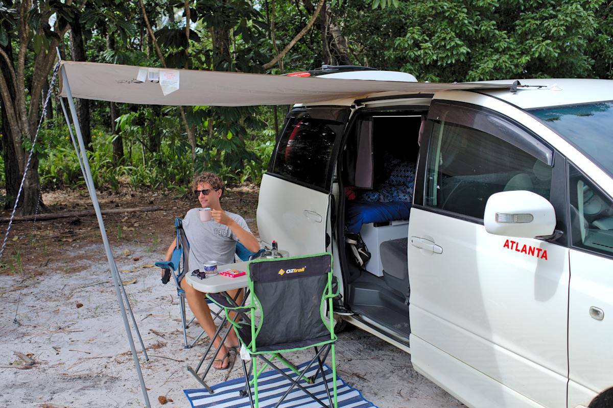 Man sitting in a camp chair at a camping table under a side awning from a campervan parked on a beach