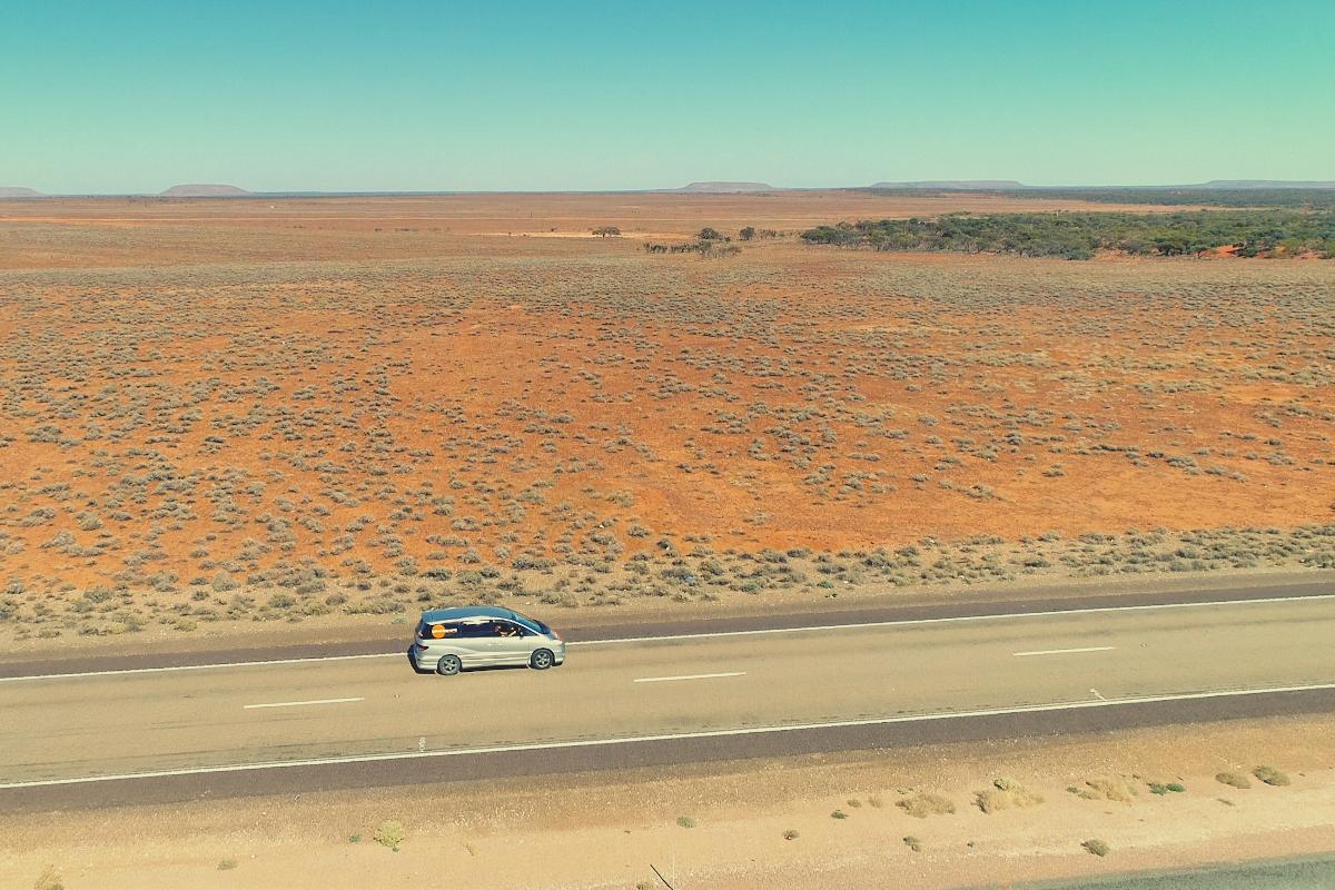 Silver campervan driving along a sealed road in the outback of Australia with red desert in the background
