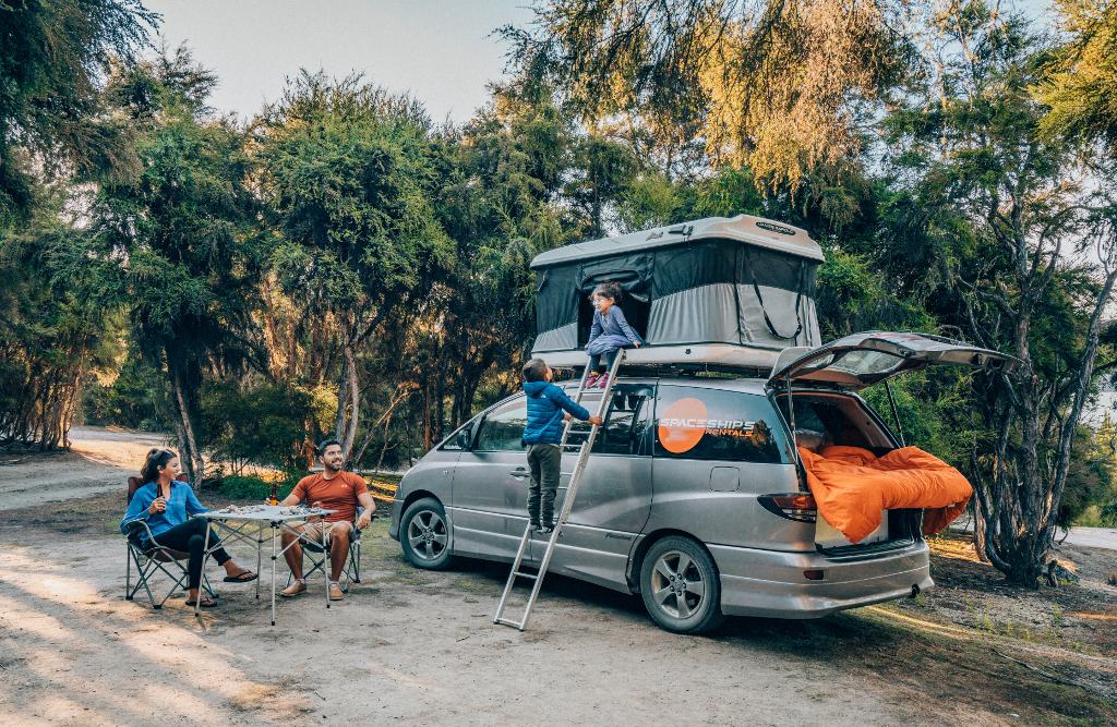 Spaceships 4-berth campervan with rooftop tent on the right, kids are sitting on the edge of the rooftop tent's bottom while their parents are sitting at the camping table next to the vehicle