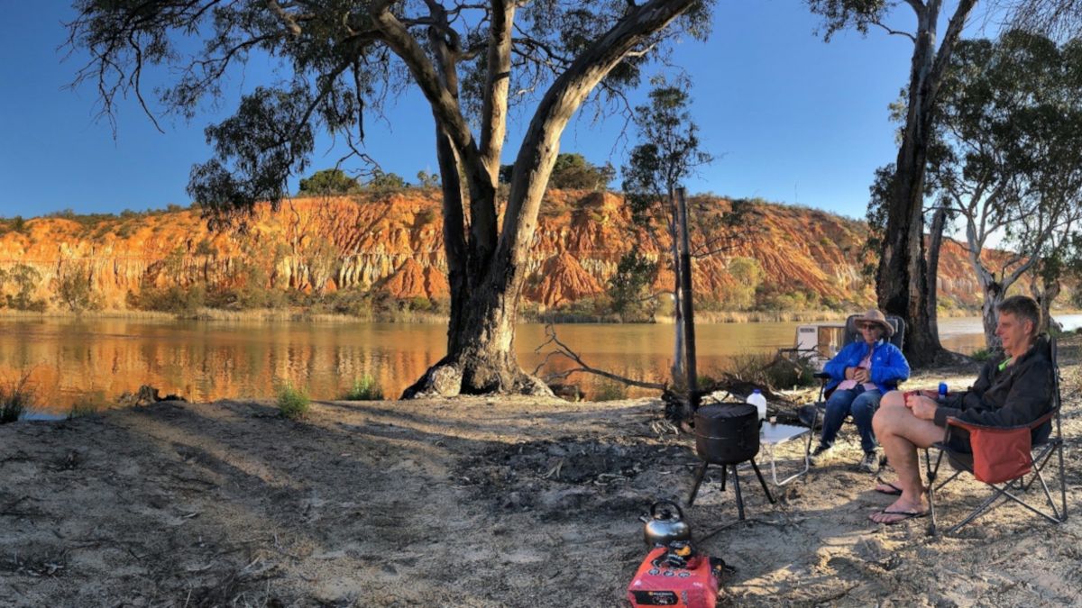 Two campers sittng on the banks of the Murray River enjoying coffee and the view. River and rock formation in the background. Trees and sandy area in the foreground