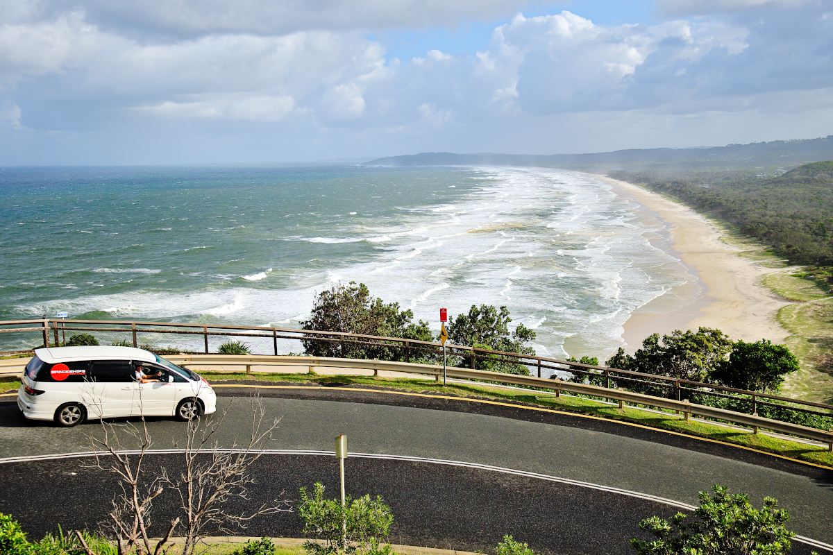 white campervan driving along a coastal road with a white sand beach streatching out in the distance